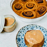 A prepared cinnamon roll with maple pecan frosting on a plate with a second cinnamon roll in the background, all placed on a table with a floral pattern. A jar of maple pecan frosting is also visible in the foreground.