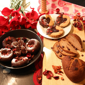 Baked goods including cookies and bread on a table with red flowers and a decorative stand.